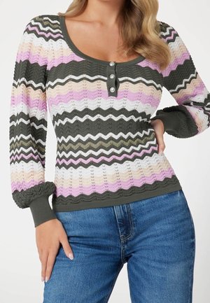 Vrouw draagt een gebreide trui met lange mouwen, voorzien van roze, witte, beige en groene zigzagstrepen en drie knopen aan de voorkant, gecombineerd met een blauwe spijkerbroek.