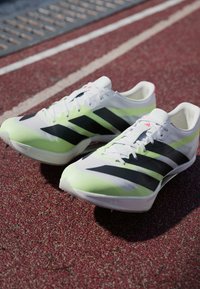 Chaussures de sport blanches avec des bandes noires et des accents vert néon. Comprend un design léger et une tige texturée, adaptées à une utilisation sur piste.