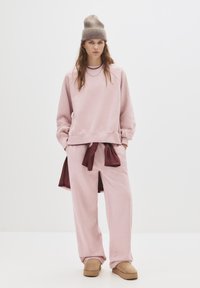 Różowy sweter z ribbowymi mankietami w połączeniu z różowymi dresami z maroonowym wiązaniem. Całość dopełnia beżowa, wełniana czapka.