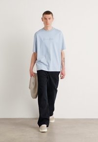 Light blue cotton t-shirt with embroidered logo, paired with dark loose-fitting pants. Model holding a beige bag and wearing cream sneakers.