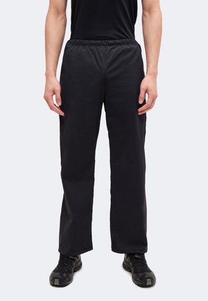 Personne portant un pantalon noir ample avec une taille élastique et des chaussures de sport noires, se tenant devant un fond uni gris clair.