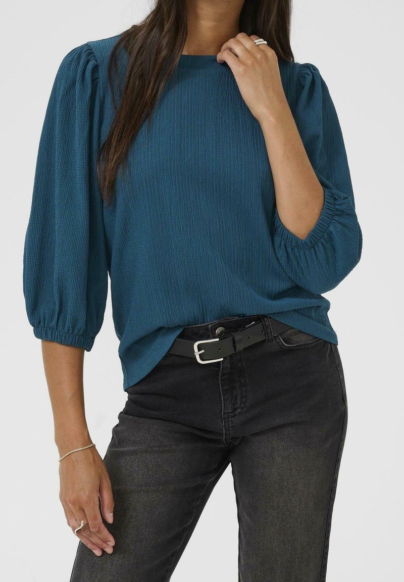 Femme portant un chemisier texturé bleu sarcelle avec des manches bouffantes trois-quarts rentrée dans un jean noir taille haute avec une ceinture noire.
