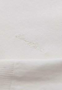 Beige brei fabric met een zachte textuur, met subtiele borduursels in een contrasterende draad. Het ontwerp bevat ribbelranddetails.