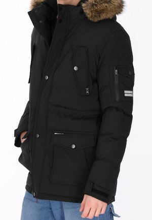 Parka d'hiver noire avec capuche bordée de fourrure, plusieurs poches zippées et boutonnées, portée avec un jean bleu, mains partiellement dans les poches.