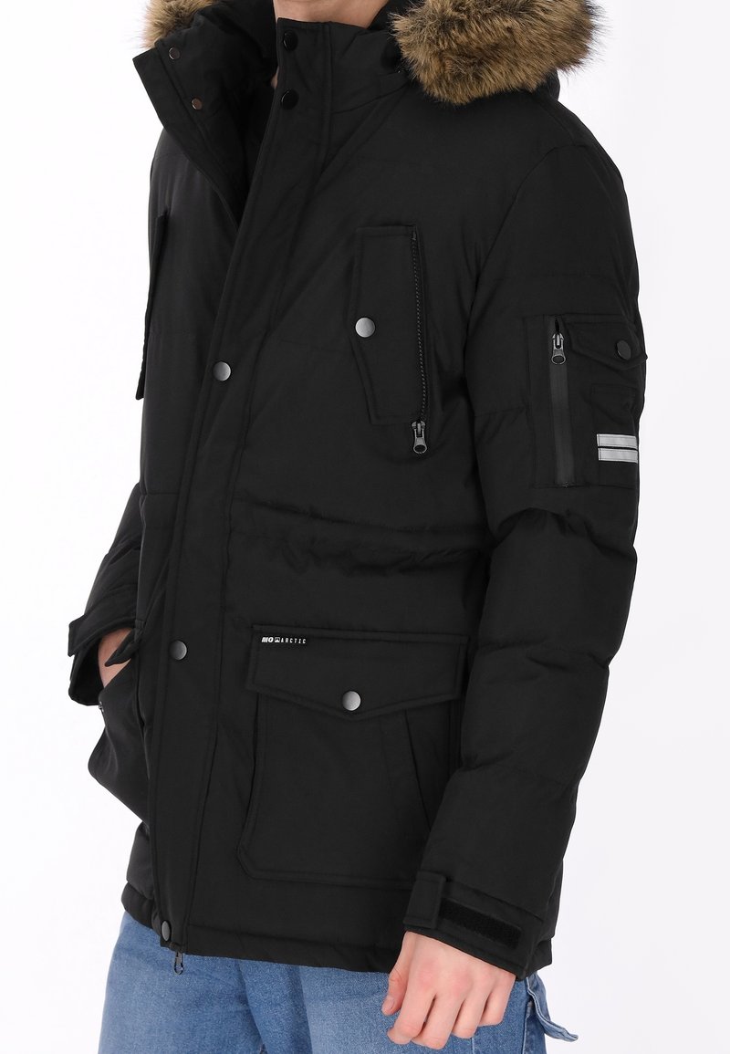 Parka d'hiver noire avec capuche bordée de fourrure, plusieurs poches zippées et boutonnées, portée avec un jean bleu, mains partiellement dans les poches.
