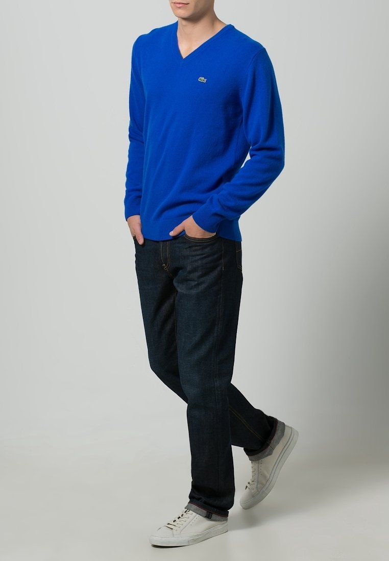 Homme portant un pull en V bleu vif, un jean foncé et des baskets blanches, debout les mains dans les poches devant un fond uni.