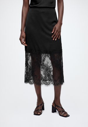 Femme portant une jupe midi noire avec un détail en dentelle florale transparente à l'ourlet et des sandales noires à talons avec des fines lanières.