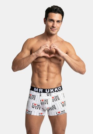 Uomo senza maglietta forma un cuore con le mani, indossa boxer bianchi con la scritta "Amo la mia fantastica moglie" e una fascia nera con la scritta "MR UKKO."