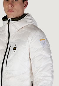Valkoinen puhvis takki, jossa on huppu, vuorattu pinta, musta vetoketju, etutaskut ja logo-patch hihassa.