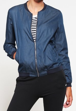 Person iført en blå denim bomberjakke over en sort og hvid stribet skjorte, kombineret med sorte bukser, stående med den ene hånd i lommen.