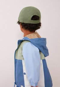 Giacca colorata in azzurro chiaro, verde e blu navy con polsini elasticizzati e un colletto a fantasia, abbinata a un cappellino verde.