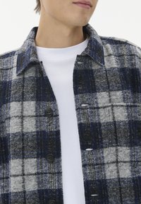 Moški nosi modro, sivo in črno karirano srajco na gumb čez enostavno belo majico s krožnim izrezom, obrnjeno rahlo levo.