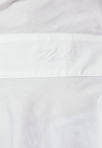 Tessuto bianco con una texture liscia e un ricamo "Karl" in filo bianco. Accentuato da una cucitura pulita e netta.