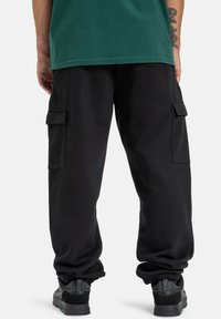 Schwarze Cargo-Hosen mit verstellbaren Bündchen, ausgestattet mit zwei Seitentaschen und einem lockeren Schnitt, kombiniert mit einem grünen Hemd.