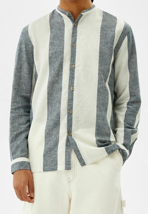 Homme portant une chemise à manches longues boutonnée avec de larges rayures verticales grises et blanches et un pantalon crème avec une main dans la poche.