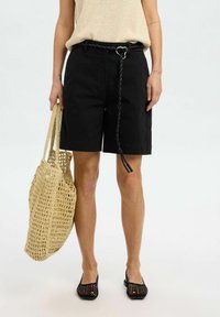 Črne bombažne kratke hlače z pletenim pasom, v kombinaciji s bež pletenim topom, velikim tkanim toaletnim nahrbtnikom in črnimi mrežastimi balerinkami.