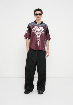 Krátke tričko bordovej a čiernej farby s ozdobným bielym grafickým vzorom, kombinované s voľnými čiernymi nohavicami s červeným prešitím a čiernymi topánkami.