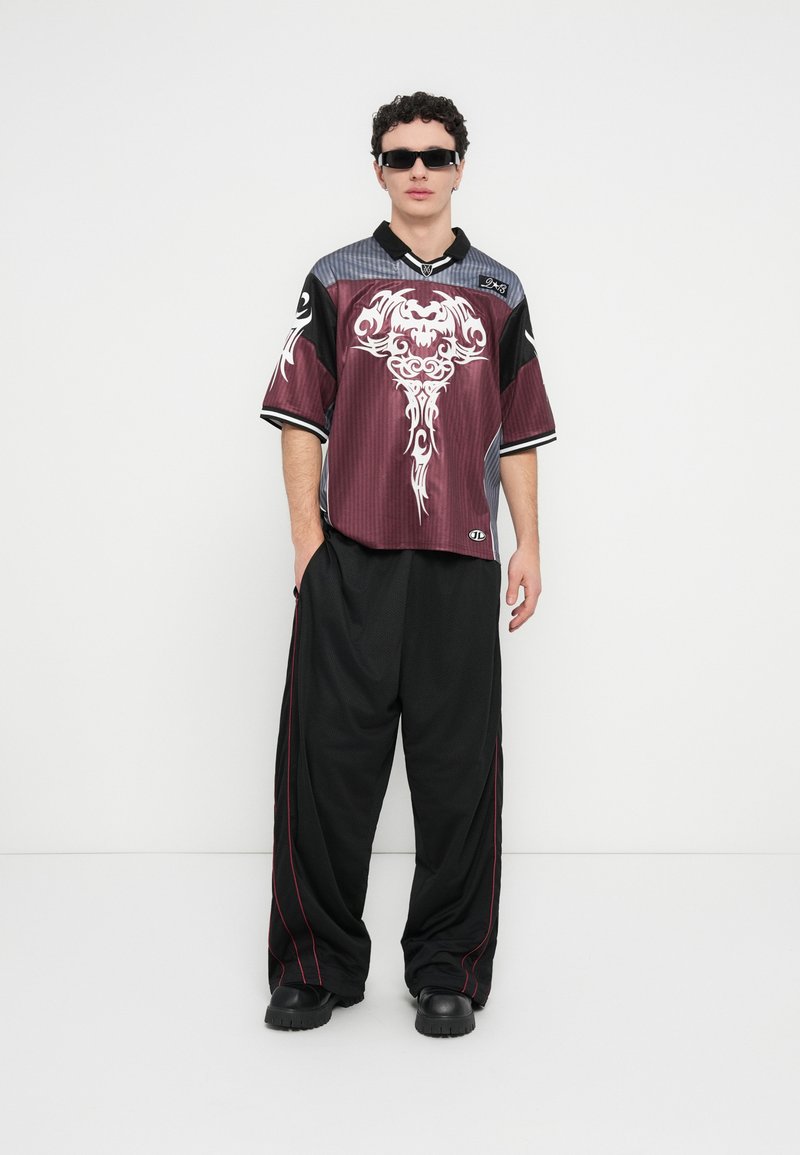 Μπλούζα με σύντομο μανίκι σε βυσσινί και μαύρο χρώμα με περίτεχνο λευκό γραφικό σχέδιο, συνδυασμένη με φαρδιά μαύρα παντελόνια με κόκκινες ραφές και μαύρα παπούτσια.