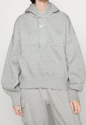 Osoba ubrana w luźną szarą bluzę z kapturem Nike ze sznurkami oraz pasujące szare spodnie dresowe z boczną kieszenią na lewej nodze.