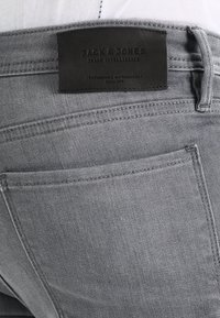 Jeans in denim grigio con etichetta in pelle nera con testo in rilievo. Sono visibili cuciture a vista e un design della tasca posteriore.
