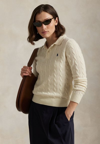 Vrouw met schildpadzonnebril, crème kabelgebreide trui, marineblauwe broek en een bruine schoudertas, poserend tegen een neutrale achtergrond.