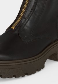 Gros plan de l'avant et du côté d'une botte en cuir marron foncé avec une semelle épaisse et une fermeture éclair dorée visible.