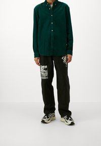 Zielona koszula ze sztruksu z kołnierzykiem na guziki, zestawiona z czarnymi spodniami ozdobionymi białymi graficznymi naszywkami. Całość dopełniają casualowe trampki.