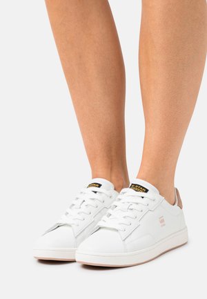 Des baskets en cuir blanc avec une texture, des lacets plats et un accent de talon beige. Présentent un logo noir et jaune sur la languette.