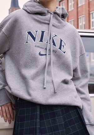 Person iført grå Nike hættetrøje med snore og navy ternet nederdel, stående ved siden af en beige bil og murstensbygninger i baggrunden.