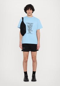 Blaues Grafik-T-Shirt mit schwarzer Grafik, schwarze Umhängetasche, schwarze Shorts und schwarze Schuhe, vor einem schlichten weißen Hintergrund.