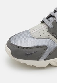 Šedé tenisky kombinující texturovanou kůži a hladkou tkaninu. Obsahují výrazné logo Nike a perforované detaily na svršku.