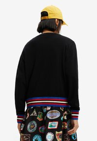 Persona che indossa un maglione nero con polsini e orlo a strisce rosse, blu e bianche, un cappello giallo e pantaloni a fantasia con toppe colorate.