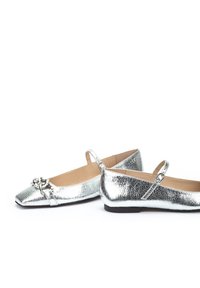 Zapatos planos metálicos plateados con punta cuadrada, que presentan un detalle decorativo de cadena y una correa en el tobillo. Cuero texturizado con un tacón bajo.