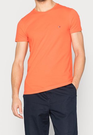 Mann trägt ein knalloranges T-Shirt mit kurzem Ärmel und Rundhalsausschnitt sowie kleinem Logo auf der Brust, kombiniert mit dunkelblauen Hosen, Hand in der Tasche.