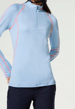 Femme portant un haut de sport à manches longues bleu clair avec des coutures orange, un pantalon noir et une sangle de casque noire autour du cou.