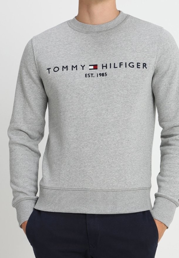 Hellgrauer Pullover mit dem Schriftzug "Tommy Hilfiger", Markenflaggenlogo und "EST. 1985" auf der Brust gestickt, getragen mit navyblauen Hosen.