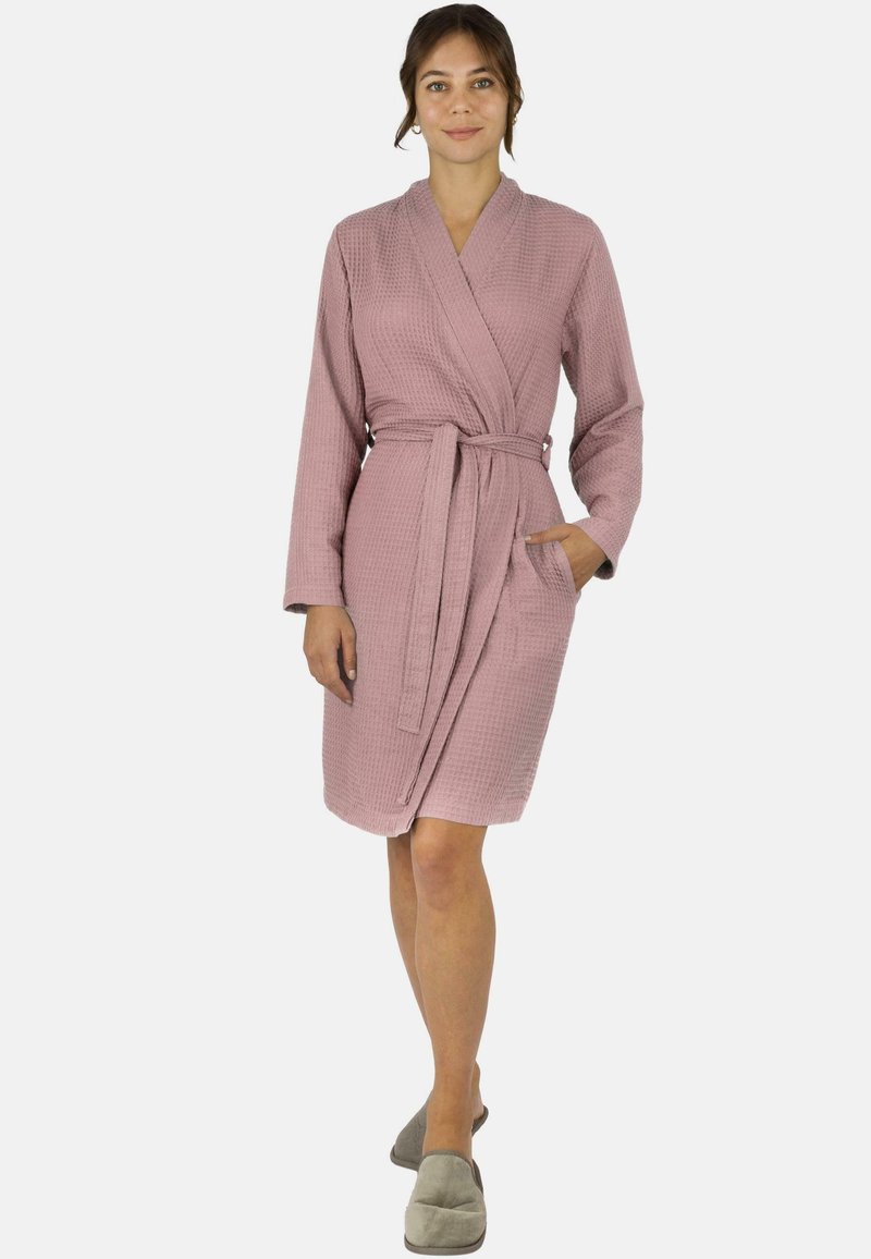 Rosa Bademantel mit Waffle-Struktur, langen Ärmeln, einem Gürtel und seitlichen Taschen. Modell trägt graue Hausschuhe.