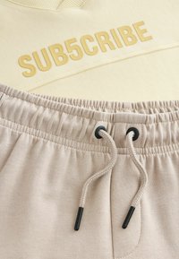 Pull jaune clair avec le texte en relief « SUB5CRIBE », associé à un pantalon de survêtement beige doté d'une taille élastique et d'un cordon de serrage.