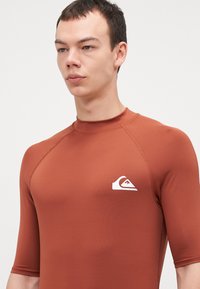 Kortermet rash guard i en rik terrakottafarge, laget av stretchstoff med en jevn tekstur. Har en hvit logo på brystet.