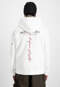 Sweatshirt à capuche blanche avec des impressions sur le devant et l'arrière. Le texte indique "Industries" et "Before Flight" en noir et rouge. Tissu doux, poignets côtelés.