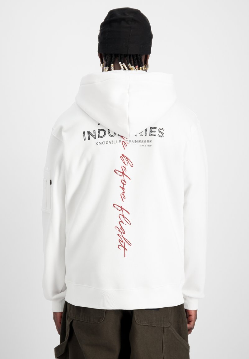Sweatshirt à capuche blanche avec des impressions sur le devant et l'arrière. Le texte indique "Industries" et "Before Flight" en noir et rouge. Tissu doux, poignets côtelés.