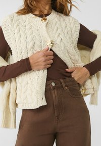 Vrouw die een crèmekleurige kabelgebreide vest met grote gouden knopen over een bruine lange mouwen top en hoge taille bruine broek draagt, terwijl ze het vest aanpast.