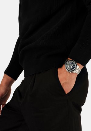 Zilveren chronograaf horloge met een zwarte wijzerplaat, metalen band en een donkere gebreide trui gecombineerd met zwarte broek, hand rustend in de zak.