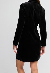 Schwarzes, samtenes Kleid mit langen Ärmeln, tailliertem Schnitt und leichtem Glanz. Verfügt über einen Kragen und endet oberhalb des Knies.