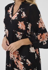 Femme portant une robe noire avec un motif floral rose et blanc et des manches cloche trois quarts, les mains délicatement jointes devant.