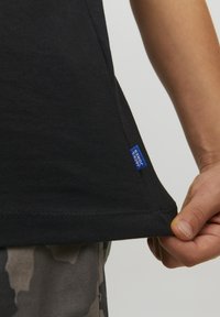 T-shirt nera in cotone. Presenta un orlo arrotondato, dettagli cuciti e una piccola etichetta blu del marchio sul lato.