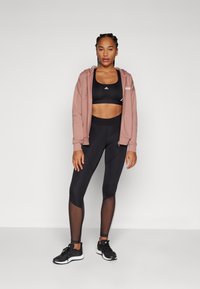 Fekete sportmelltartó, rózsaszín cipzáras kapucnis felső és fekete leggings, hálós díszítéssel; atlétikai cipők egészítik ki az outfite-t. Egyszerű és funkcionális dizájn.