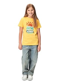 Gul bomulds t-shirt med grafisk print af sol, bølger og teksten "sommer vanvid." Kombineret med lyseblå denimjeans og hvide sneakers.