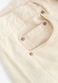 Lichtbeige broek van een zachte, gestructureerde stof met een klein achterzakje dat is beveiligd met zilveren hardware. Subtiele diagonale strepen zichtbaar.