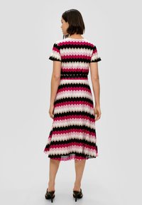 Gestreiftes Kleid mit einem Zickzackmuster in Pink, Weiß und Schwarz, mit kurzen Ärmeln und einem ausgestellten Saum. Trägt dazu hohe Schuhe.